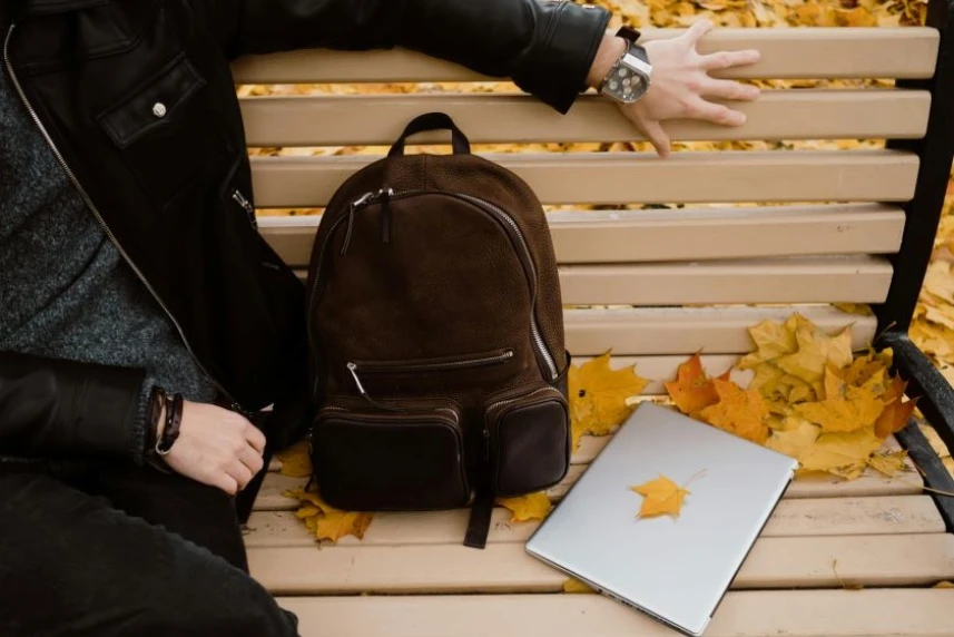 Osoba siedząca na ławce w parku. Obok znajduje się brązowy plecak i laptop. Osoba siedząca na ławce w parku. Obok znajduje się brązowy plecak i laptop.