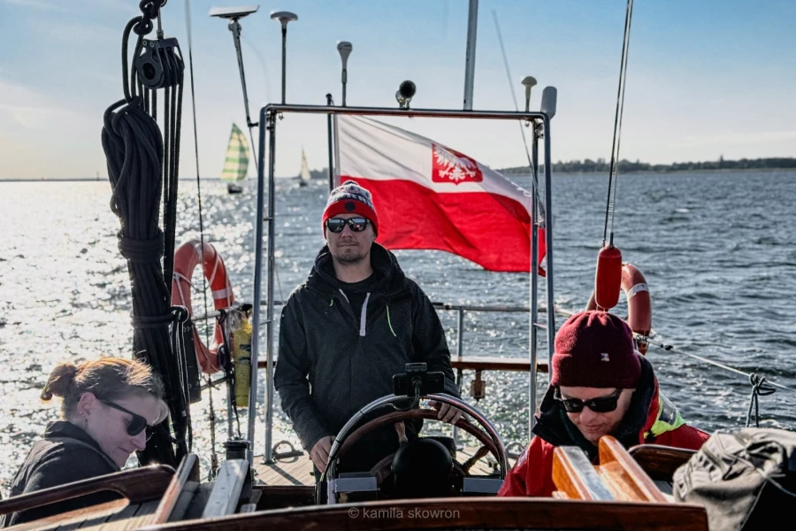 Grupa żeglarzy na akwenie wodnym. W tle biało-czerwona bandera.