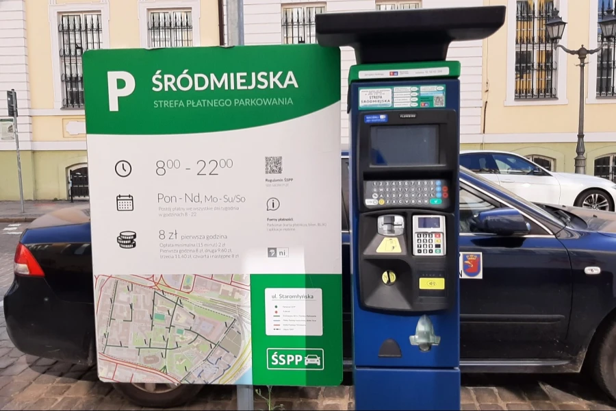 Parkomat i tablica informacyjna na tle zaparkowanych samochodów.