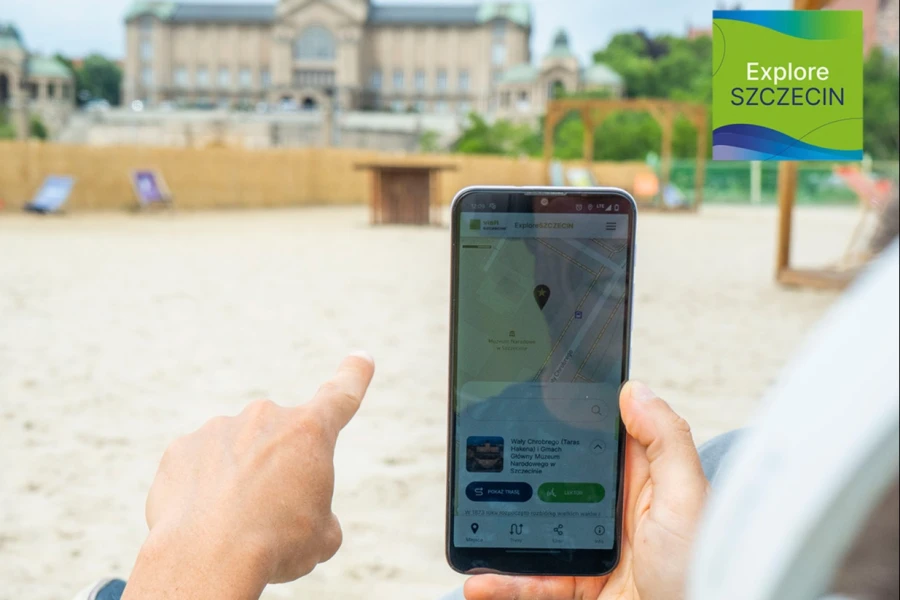 Smartfon trzymany w ręku przez osobę siedzącą na plaży miejskiej.
