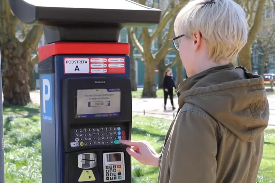 Osoba ubrana w kurtkę z kapturem przy parkomacie.